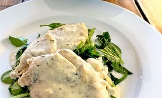 Two pieces of chicken breast in a creamy white herb sauce served on a bed of fresh spinach leaves on a white plate, with a bowl of whole baby potatoes in the background.