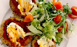 Three breaded aubergine slices topped with tomato sauce and melted cheese are served on a white plate with a side salad of mixed leaves and cherry tomatoes.