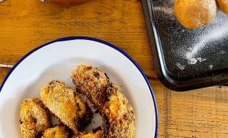 A plate of crispy breaded chicken pieces, a saucepan of green beans, and a baking tray with baked potatoes on a wooden table.