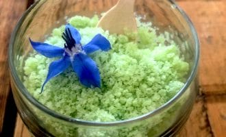 A glass beaker filled with light green granulated pudding, topped with a vibrant blue edible flower, and a small wooden spoon resting inside. The beaker sits on a rustic wooden table.