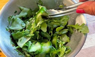 A hand with red nail varnish holds tongs, tossing a green salad with cucumber ribbons and leafy greens in a metal bowl on a wooden surface.