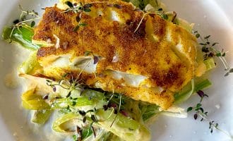 A golden-brown, pan-seared fish fillet sits atop a creamy medley of sautéed vegetables, garnished with fresh microgreens, on a white plate.