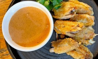 A black plate with crispy chicken wings, a garnish of coriander, and a white bowl filled with brown dipping sauce, set on a wooden table.