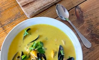 A white bowl of yellow seafood soup garnished with coriander sits on a wooden table next to a metal spoon and a chopping board with a piece of crusty bread. Black mussel shells are visible in the soup.