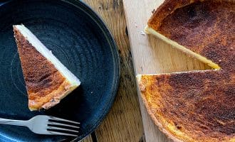 A slice of custard tart on a black plate with a fork beside it sits next to the rest of the tart, which is on a wooden chopping board with one slice missing. The surface of the tart appears caramelised.