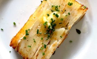 A rectangular slice of layered, golden-brown potato garnished with chopped chives and a pat of melted butter, served on a white plate.