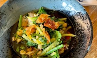 A bowl of vegetable soup garnished with fresh herbs and crispy toppings sits on a wooden table, with a decorative spoon and glass visible nearby.