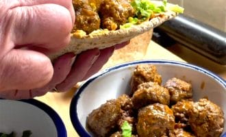 A hand holds a pitta filled with meatballs, lettuce, and sauce. Below, a bowl contains more saucy meatballs, and a small bowl of chopped herbs is nearby on a wooden worktop.