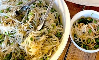 A large white bowl of noodle salad with green vegetables and metal tongs sits on a wooden table next to a smaller bowl of salad and a plate of sliced beef over greens.