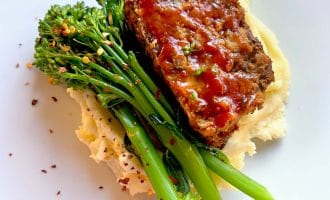 A slice of glazed meatloaf sits on creamy mashed potatoes, garnished with steamed tenderstem broccoli and sprinkled with red chilli flakes, served on a white plate.
