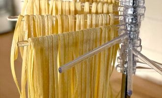 Freshly made pasta noodles hanging to dry on a clear plastic drying rack in a kitchen setting. The uncooked pasta strands are neatly arranged in rows.
