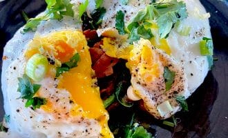Two fried eggs with runny yolks are garnished with fresh herbs and chopped spring onions, served on a dark plate. A fork cuts into one egg, letting the yolk flow out over sautéed vegetables underneath.