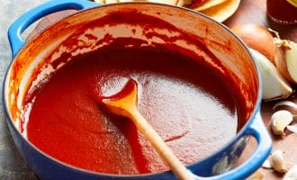 A blue pot filled with smooth red enchilada sauce sits on a wooden surface, surrounded by a wooden spoon, dried chillies, corn tortillas, a halved onion, garlic cloves, and a jar of hot sauce.