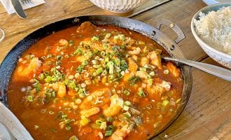 A frying pan filled with a red tomato-based stew topped with chopped spring onions sits on a wooden table, alongside a bowl of white rice, a salad, and empty plates with cutlery.