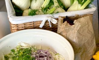 A wicker basket filled with fresh fennel bulbs sits behind a bowl containing sliced fennel, red onion, and herbs. A brown paper bag and two lemons are also on the table.