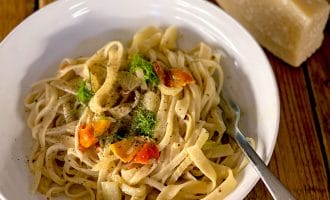 A white bowl of fettuccine pasta with cherry tomatoes, fresh herbs, and black pepper. A fork rests on the bowl, and a block of cheese with a grater is in the background on a wooden table.