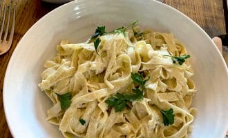 A bowl of fettuccine Alfredo topped with fresh parsley sits on a wooden table next to a bowl of chopped herbs, a glass of wine, and a fork.