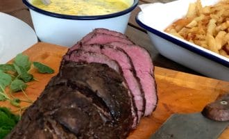 Sliced roast beef on a wooden chopping board with a knife, next to a bowl of chips and a bowl of yellow sauce with herbs, all set on an outdoor wooden table.