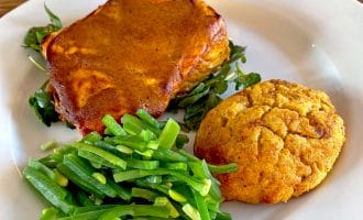 A white plate with a serving of lasagne on leafy greens, a scoop of sautéed green beans, and a golden-brown scone. The food is arranged neatly on a wooden table.