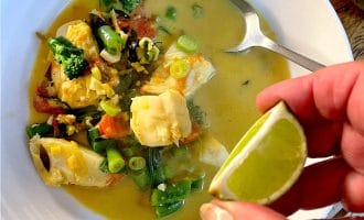 A hand squeezes a lime wedge over a bowl of fish and vegetable stew in a yellow broth, with a spoon resting inside the white bowl.