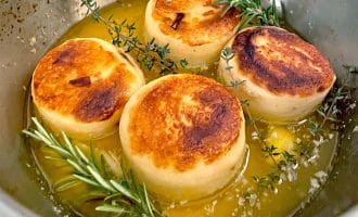 Four golden-brown, round potato slices are seared in a pan with melted butter, garnished with sprigs of fresh rosemary and thyme.
