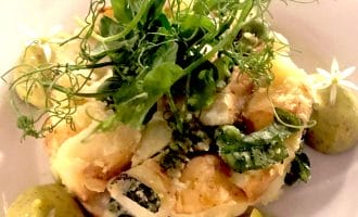 A plated dish featuring a creamy vegetable medley garnished with fresh herbs and edible flowers, accompanied by three dollops of green sauce, each topped with a small white flower.