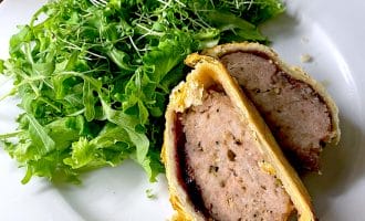 A plate with a leafy green salad and a sliced portion of meat wrapped in golden puff pastry, resembling a homemade pork Wellington, on a white plate.