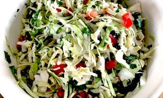 A white bowl filled with shredded cabbage salad mixed with chopped tomatoes, coriander, and green herbs, placed on a wooden surface.
