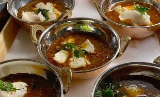 Six metal bowls filled with spicy red curry, garnished with herbs and chunks of white fish, are arranged on a tray. The curry has a rich, oily surface and each bowl has ornate metal handles.