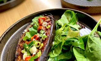 A dark plate with a roasted aubergine half topped with chopped spring onions, red chilli, and herbs, served alongside a fresh green salad.