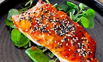 A fillet of salmon topped with a red glaze and garnished with white and black sesame seeds, served on a bed of fresh green leaves on a dark plate.