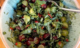 A bowl containing chopped green herbs, red and green grapes, sliced red chilli, and black olives, with a spoon resting in the mixture on a wooden surface.