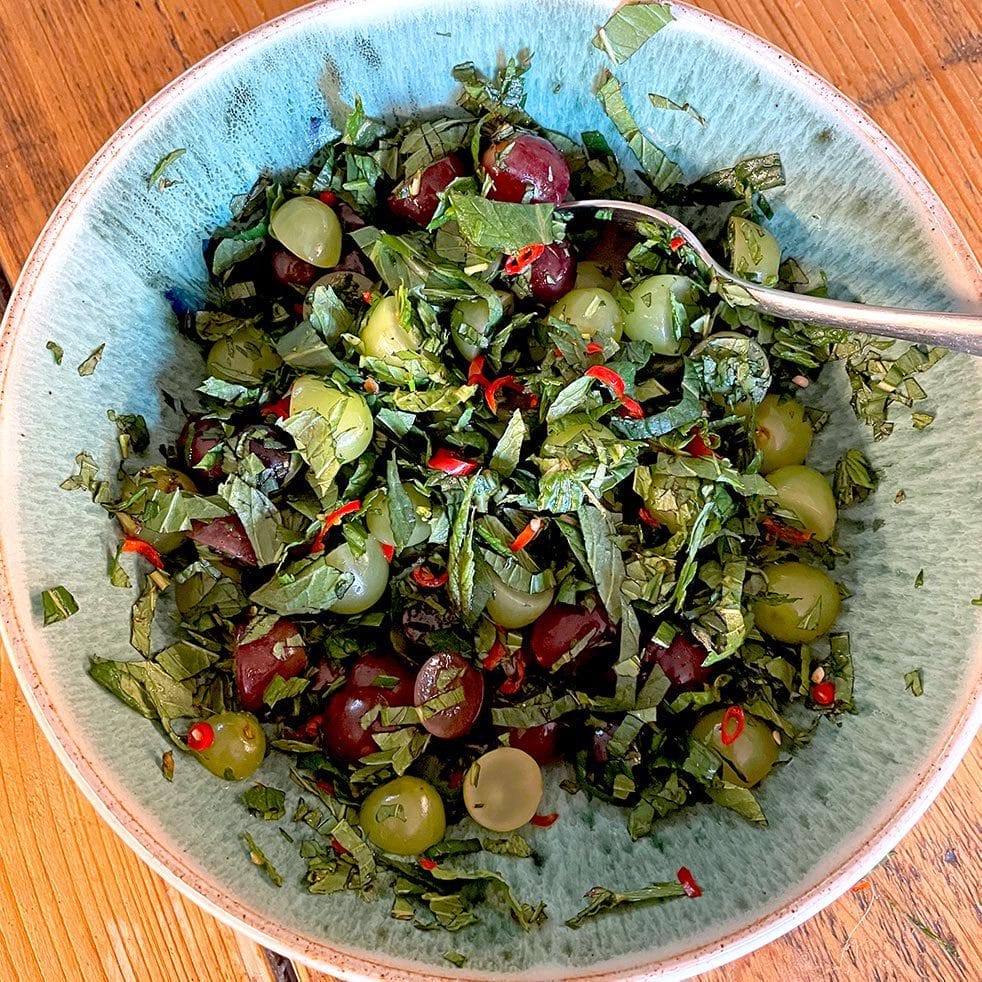 A bowl containing chopped green herbs, red and green grapes, sliced red chilli, and black olives, with a spoon resting in the mixture on a wooden surface.