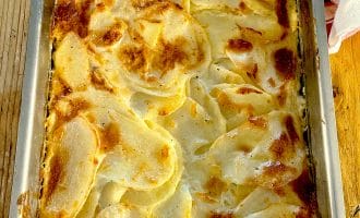 A baked potato gratin in a rectangular metal dish, featuring thinly sliced potatoes layered with cream and cheese, with a golden-brown, slightly crispy top. The dish sits on a wooden surface.