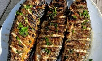 Three grilled whole fish with char marks and garnished with chopped herbs are served on a white oval plate, set on a rustic wooden table in bright sunlight.