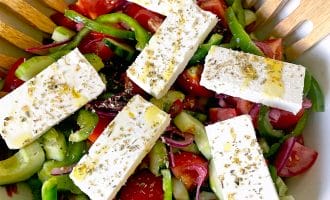 A bowl of Greek salad with sliced tomatoes, cucumbers, green peppers, red onions, and five rectangular pieces of feta cheese, garnished with dried herbs and olive oil. Wooden salad servers rest at the side.