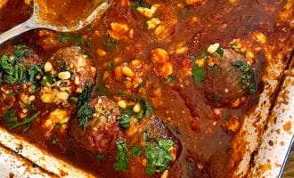 A baking dish filled with meatballs in a rich tomato sauce, topped with crumbled cheese, chopped herbs, and pine nuts. Two metal serving spoons rest in the dish.