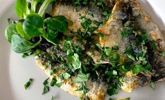 Grilled fish fillets garnished with chopped herbs, served with a wedge of lemon and a small side of fresh salad leaves on a white plate.