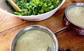A bowl of chopped green salad with tongs next to two bowls of creamy green herb dressing on a wooden table.