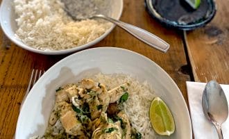 A bowl of white rice topped with chicken, vegetables, and herbs sits on a wooden table next to a lime wedge, spoon, serviette, plate with another lime wedge, and a bowl of plain rice with a spoon.