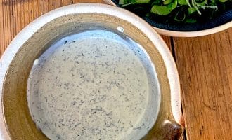 A rustic ceramic bowl filled with creamy herb dressing sits on a wooden table next to a dark bowl of fresh rocket and cucumber salad.