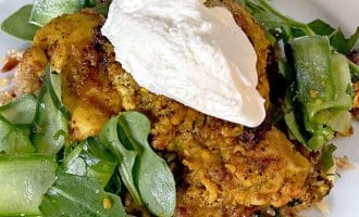 Seasoned roasted chicken topped with a dollop of creamy white sauce, served on a bed of rocket leaves and cucumber ribbons, garnished with chopped nuts on a white plate.