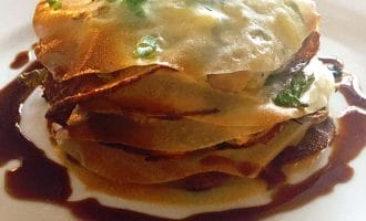 A plated dish featuring a layered savoury pastry with a golden-brown top, garnished with chopped spring onions and surrounded by a dark sauce on a white plate.