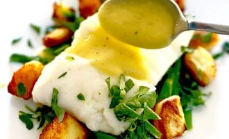 A spoon drizzles sauce over a piece of white fish on a bed of green beans and roast potatoes, garnished with fresh chopped herbs, served on a white plate.