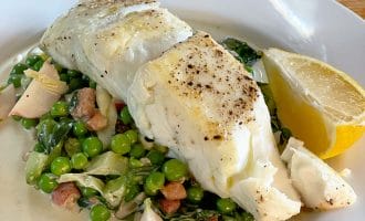 A white plate with two pieces of seasoned, cooked white fish on a bed of green peas, leafy greens, and diced vegetables, garnished with a lemon wedge and served with a light sauce.
