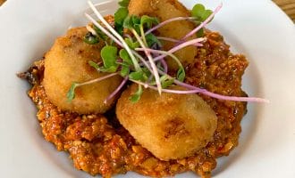 Three golden brown, breadcrumbed croquettes served on a bed of chunky red sauce, garnished with fresh green microgreens, on a white plate set on a wooden table.