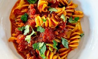 A white bowl filled with fusilli pasta topped with chunky tomato sauce and garnished with fresh parsley leaves, placed on a wooden surface.