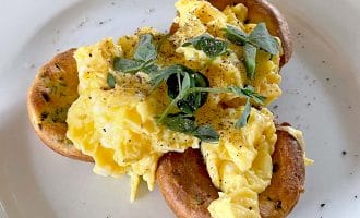 Scrambled eggs served on two toasted crumpets, garnished with fresh herbs and cracked black pepper, on a white plate.