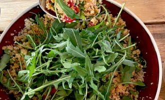 A bowl of couscous salad with chopped vegetables, garnished with fresh rocket leaves, sits on a wooden table. A spoon is serving a portion of the salad from the bowl.