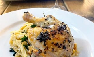 A roasted chicken leg, seasoned with herbs, is served on a bed of fettuccine pasta with greens on a white plate. In the background, a tray with more chicken and a glass of wine can be seen.